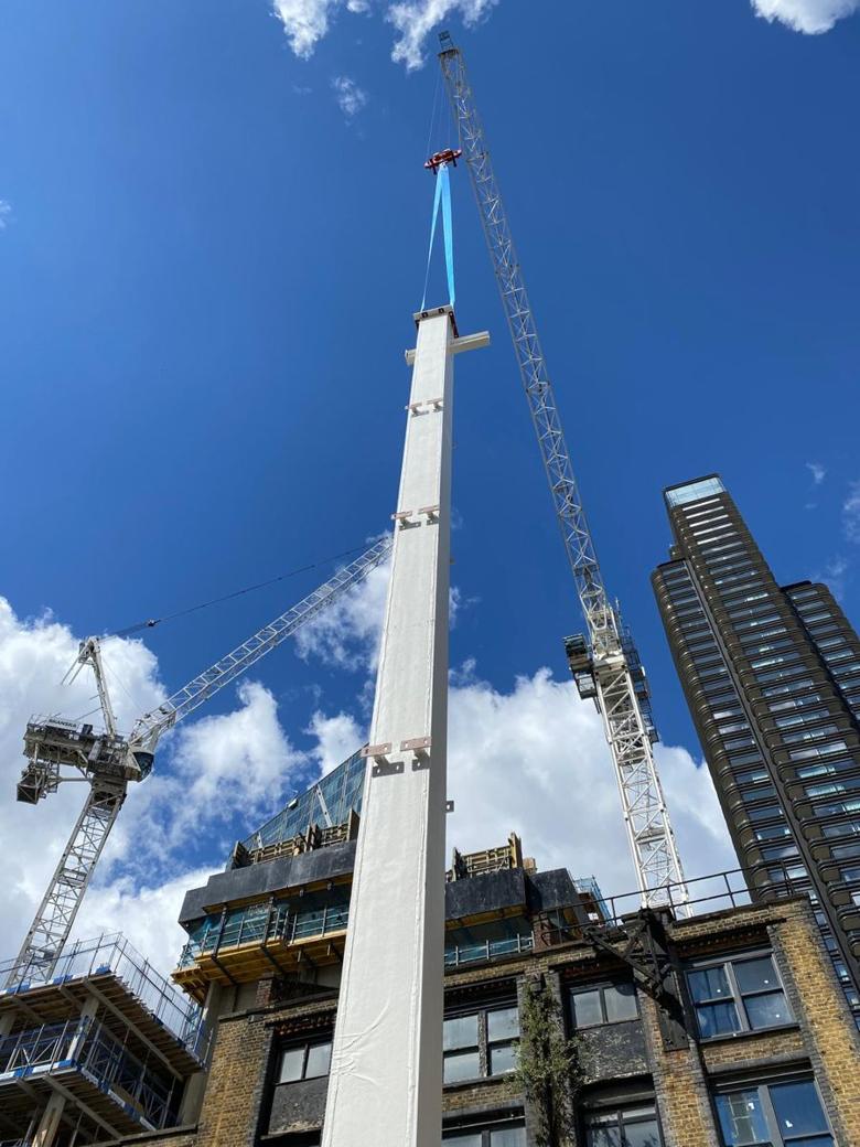 Tower crane lifting a tall white steel column on a round sling between city buildings